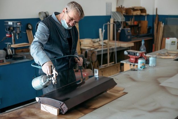 Un homme à l'âge de la retraite peint des meubles en bois dans un atelier de menuiserie avec un pistolet compresseur