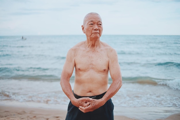 Un homme âgé sans chemise montre ses muscles alors qu'il se tient sur la plage contre le ciel au coucher du soleil.