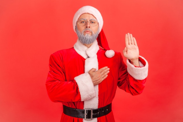 Homme âgé sérieux avec barbe grise portant le costume de père Noël debout levant la main et disant