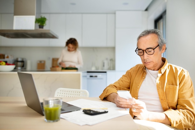 Homme âgé sérieux utilisant un téléphone portable assis à table devant un ordinateur portable dans la cuisine pendant que sa femme