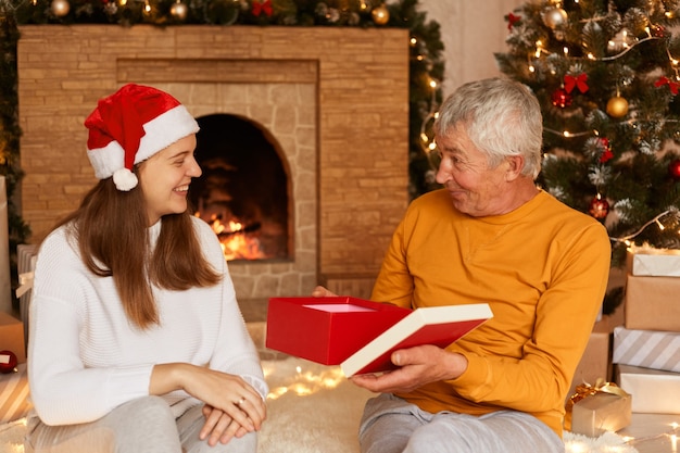 Un homme âgé surpris portant un pull orange ouvrant la boîte à cadeaux de sa fille, exprimant son étonnement, posant près de l'arbre de Noël et de la cheminée, passant le réveillon du nouvel an ensemble.