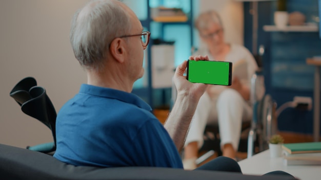 Homme âgé utilisant un écran vert horizontal sur un téléphone portable, ayant des béquilles sur un canapé. Personne retraitée détenant un modèle de clé chroma avec espace de copie isolé et arrière-plan maquette vierge sur l'appareil.