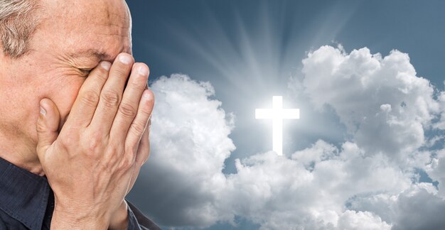 Homme âgé avec un visage fermé par les mains sur le fond du ciel avec une croix, symbole de la foi