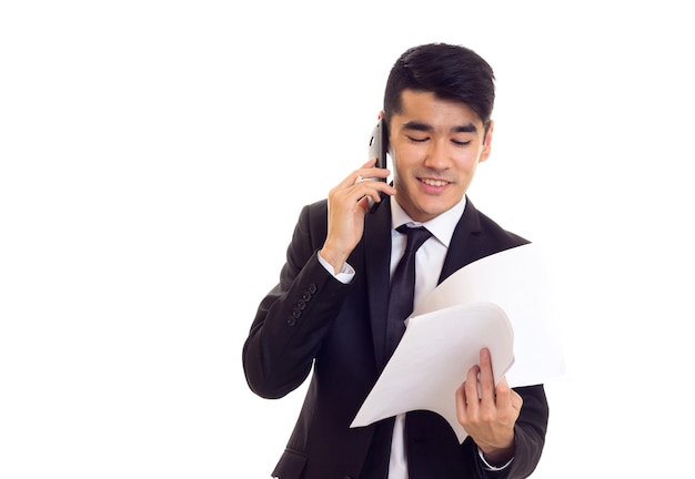 Homme agréable aux cheveux noirs en chemise blanche et costume noir avec cravate tenant des papiers et parlant au téléphone sur fond blanc
