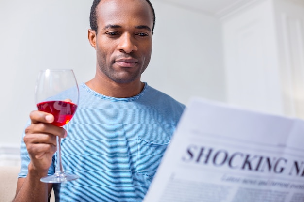 Homme agréable joyeux positif assis à la maison et avoir du vin en lisant des articles