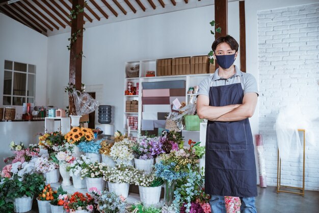 Homme à l'aide de masque facial fleuriste debout bras croisé avec des fleurs fraîches dans sa boutique de fleurs suivant protocole sain