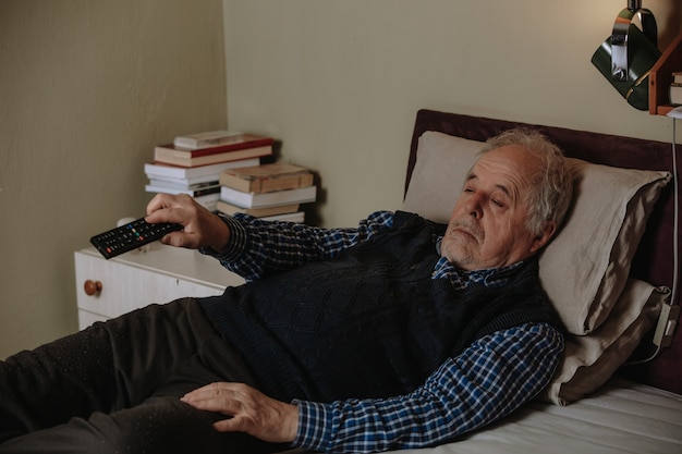 Homme aîné faisant la sieste en position couchée dans son lit et regardant la télévision avec une télécommande de télévision, se reposant dans un lit