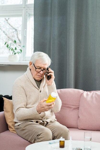 Homme aîné avec un téléphone portable assis sur un canapé, regardant une bouteille de pilules dans ses mains et consultant son médecin sur la façon de les prendre