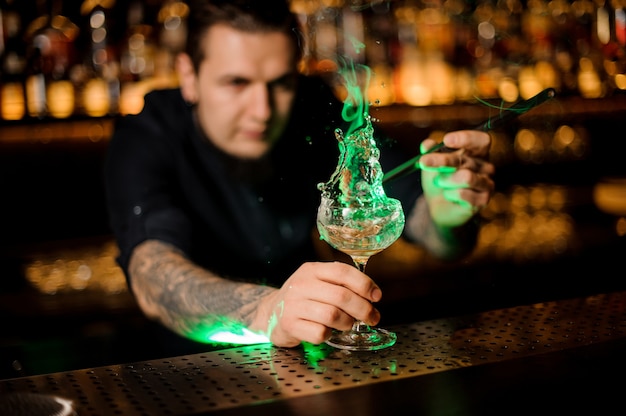 Homme ajoutant au verre à cocktail avec une cannelle fumée aromatique orange séchée avec des pincettes sur le comptoir du bar