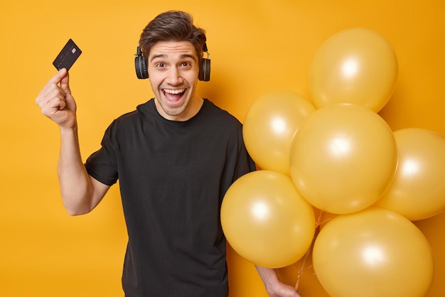 Un homme d'anniversaire positif tient une carte de crédit et un tas de ballons gonflés prêts pour une occasion spéciale de célébration écoute de la musique via un casque stéréo avec un son fort isolé sur fond jaune