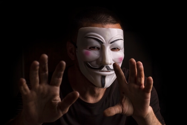 Homme anonyme portant un masque de Guy Fawkes avec les mains levées dans une pièce sombre