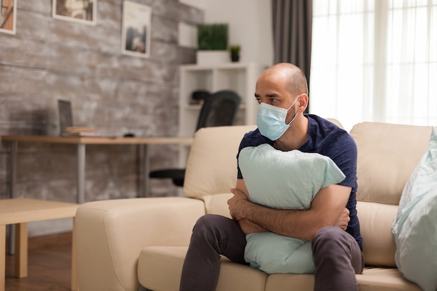 Homme anxieux tenant fermement un oreiller pendant l'auto-isolement portant un masque jetable.