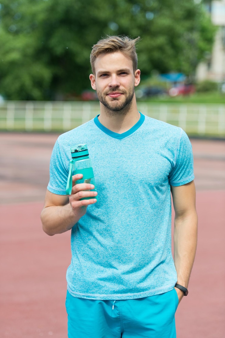 Photo un homme à l'apparence athlétique tient une bouteille d'eau. l'athlète boit de l'eau après l'entraînement au stade par une journée ensoleillée. athlète homme en vêtements de sport s'entraînant en plein air. concept de sport et de mode de vie sain.