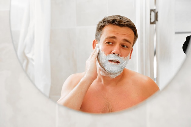 Un homme applique de la mousse à raser sur son visage et se regarde dans un miroir rond