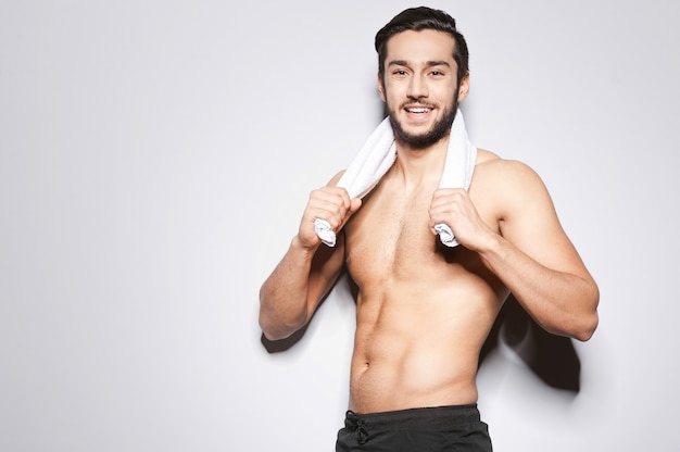 Homme après entraînement. Beau jeune homme avec une serviette sur les épaules regardant la caméra et souriant en se tenant debout sur fond gris