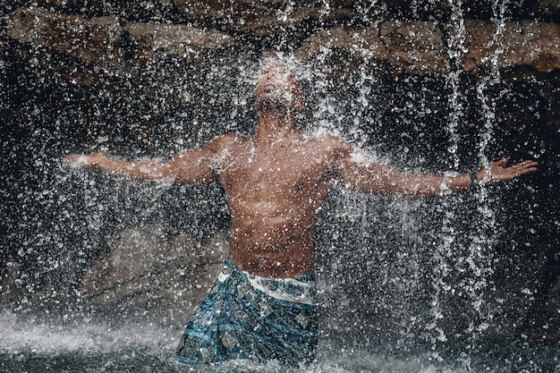 Homme arabe aux seins nus en forme et beau sous la cascade, d'énormes rochers et de l'eau, des éclaboussures et des taches; concept de beauté naturelle.