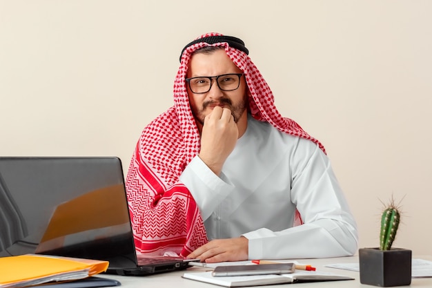 Un homme arabe, un homme d'affaires, un cheikh en costume national arabe travaille à une table au bureau. Investissements, affaires, travail via Internet, contrats en ligne.