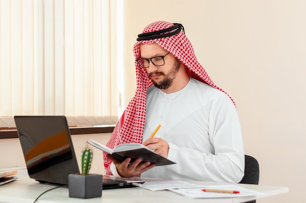 Un homme arabe, un homme d'affaires, un cheikh travaille sur un ordinateur portable. Investissements, affaires, travail via Internet, contrats en ligne.