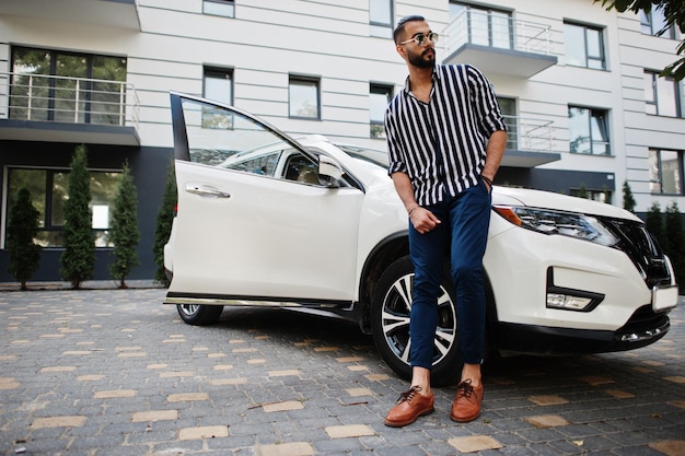 Un homme arabe réussi porte une chemise rayée et des lunettes de soleil pose près de sa voiture suv blanche. Hommes arabes élégants dans les transports.