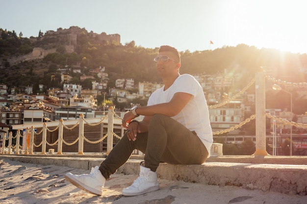 Un homme arabe riche en mode porte des lunettes de soleil et un t-shirt blanc posé contre la mer d'Oman.