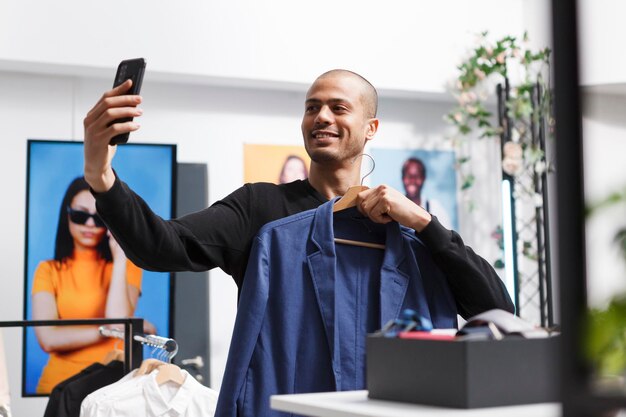 Homme arabe souriant tenant un cintre avec une veste élégante tout en prenant un selfie à l'aide de la caméra frontale du smartphone dans un magasin de vêtements. Blogger présentant des vêtements aux abonnés des réseaux sociaux dans la boutique