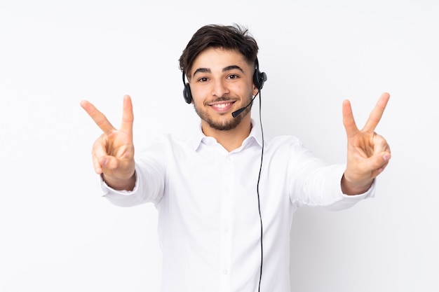 Homme arabe télévendeur travaillant avec un casque isolé sur un mur blanc souriant et montrant le signe de la victoire