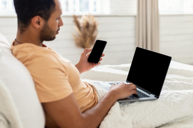 Homme arabe tenant un téléphone portable et un ordinateur portable assis dans son lit à l'intérieur