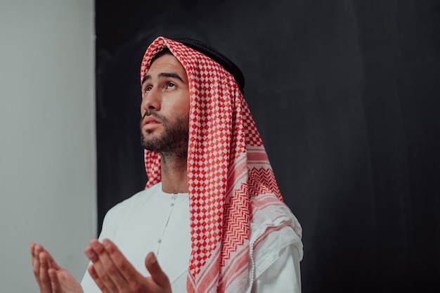 Un homme arabe en vêtements traditionnels faisant la prière traditionnelle à Dieu, garde les mains dans un geste de prière devant un tableau noir représentant la mode moderne de l'islam et le concept de ramadan kareem.