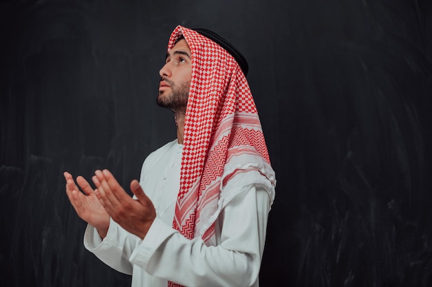 Un homme arabe en vêtements traditionnels faisant la prière traditionnelle à Dieu, garde les mains dans un geste de prière devant un tableau noir représentant la mode moderne de l'islam et le concept de ramadan kareem.