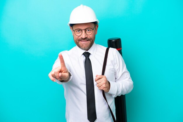 Homme d'architecte d'âge moyen avec casque et tenant des plans isolés sur fond bleu montrant et levant un doigt