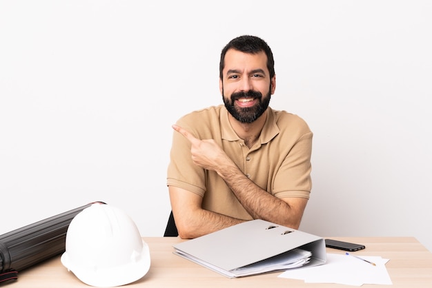 Homme architecte caucasien avec barbe dans une table pointant vers le côté pour présenter un produit.