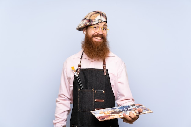 Homme artiste rousse avec une longue barbe tenant une palette sur un mur bleu isolé avec une expression heureuse