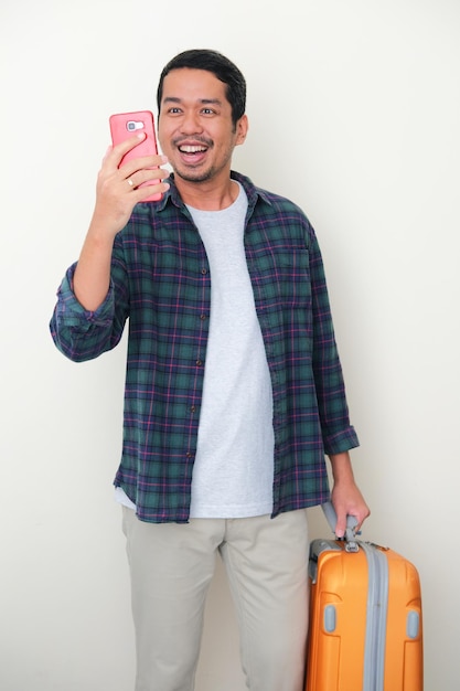 Un homme asiatique adulte apporte une valise de voyage montrant une expression heureuse en regardant son téléphone portable