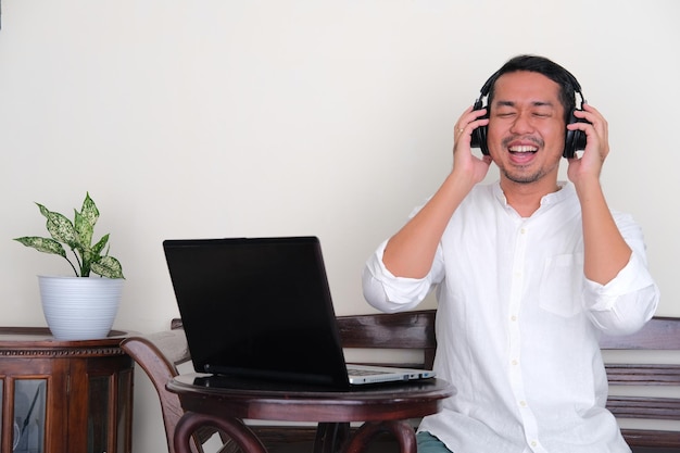 Homme asiatique adulte appréciant la musique avec une expression heureuse assis devant un ordinateur portable