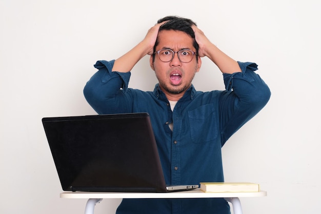 Un homme asiatique adulte assis devant son ordinateur portable montrant une expression de visage choquée