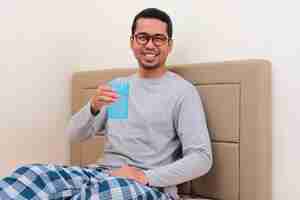 Photo homme asiatique adulte assis sur son lit souriant tout en tenant un verre d'eau