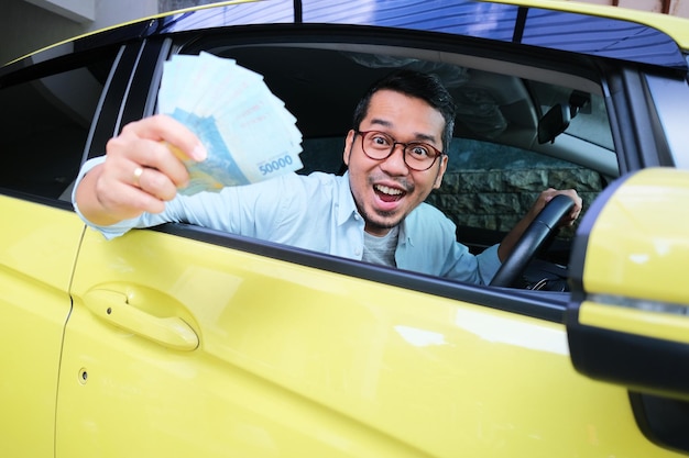 Homme asiatique adulte montrant de l'argent avec une expression excitée de l'intérieur de la fenêtre de sa voiture