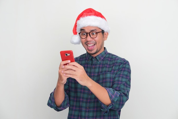 Homme asiatique adulte portant un chapeau de Noël regardant son téléphone portable avec une expression excitée