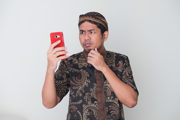 Homme asiatique adulte portant un costume javanais montrant une expression confuse en regardant son téléphone portable