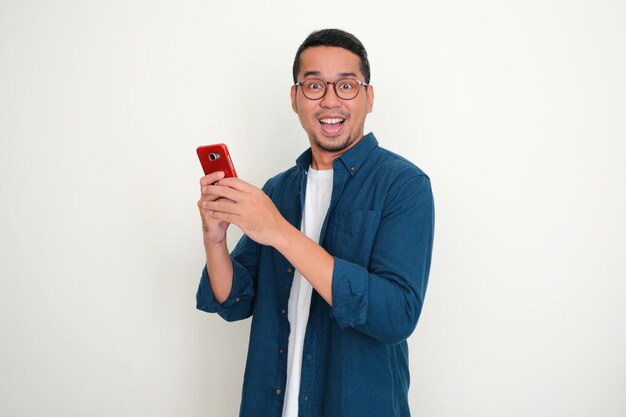 Homme asiatique adulte regardant la caméra avec une expression heureuse tout en utilisant son téléphone