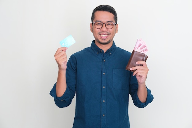 Homme asiatique adulte souriant confiant tout en tenant un portefeuille plein d'argent et une carte de crédit vierge