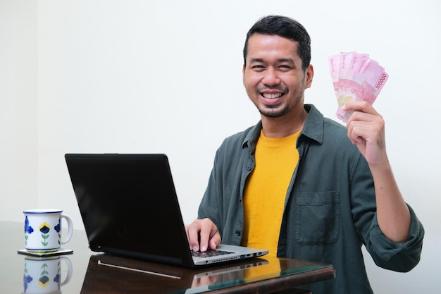 Homme asiatique adulte souriant devant son ordinateur portable tout en tenant de l'argent