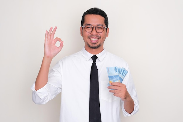 Homme asiatique adulte souriant et faisant signe du doigt OK tout en tenant de l'argent