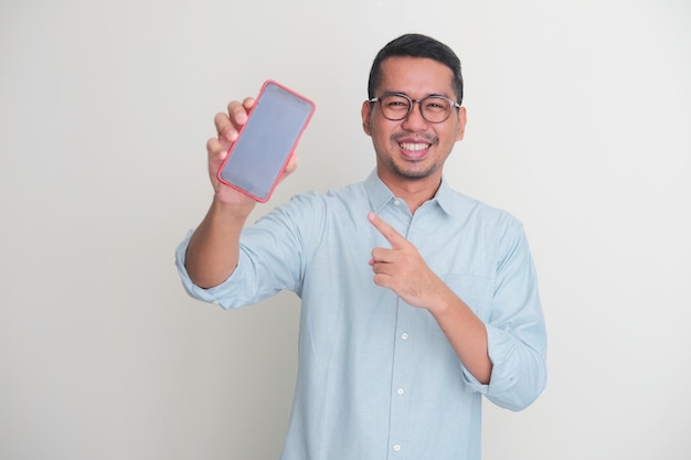 Photo homme asiatique adulte souriant heureux tout en montrant un écran de téléphone portable vierge et en pointant dessus