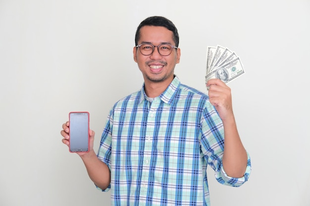 Homme asiatique adulte souriant heureux tout en tenant la monnaie du dollar américain et en montrant l'écran du téléphone portable vide