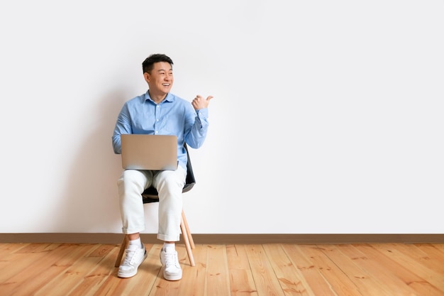 Homme asiatique d'âge moyen excité assis sur une chaise avec un ordinateur portable et pointant de côté l'espace libre sur le mur blanc du studio