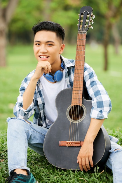 Homme asiatique assis sur l'herbe verte avec guitare et souriant à la caméra à l'extérieur