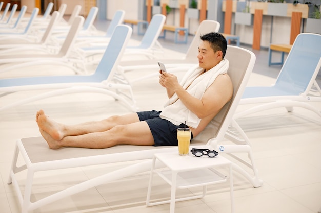 Homme asiatique assis sur un lit de bronzage dans une piscine intérieure et utilisant un téléphone