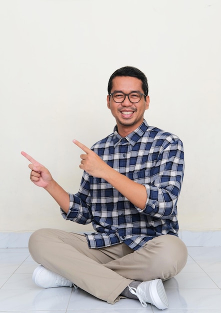 Photo homme asiatique assis sur le sol souriant à la caméra tout en pointant vers le côté droit