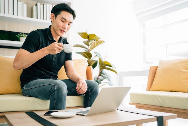 Homme asiatique assis et travaillant sur le canapé en buvant du café. Homme d'affaires occasionnel travaillant sur ordinateur portable à la maison. Homme travaillant dans une zone de détente au bureau. Période de quarantaine commerciale.